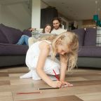 Cute daughter drawing with colored pencil sitting on warm house floor while parents using laptop on sofa, creative little kid girl playing alone, family leisure activities and hobbies at home concept